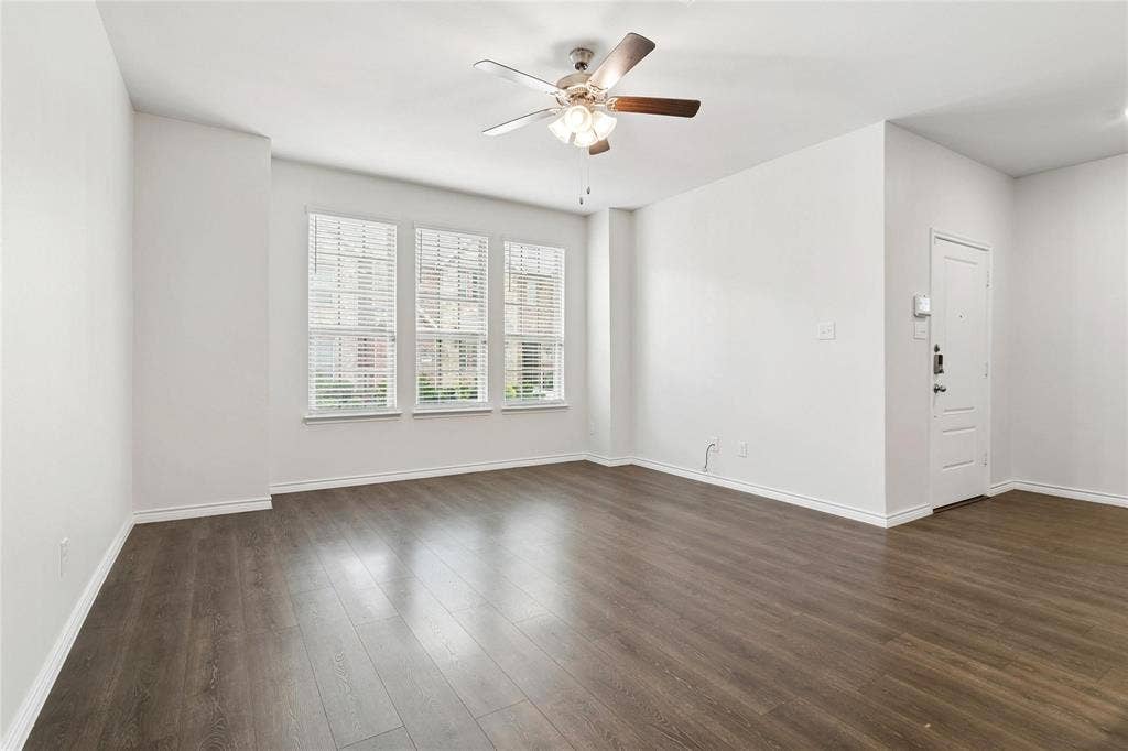 Living space featuring wood-finish flooring, a multi-blade ceiling fan with integrated lighting, and three tall windows with horizontal blinds