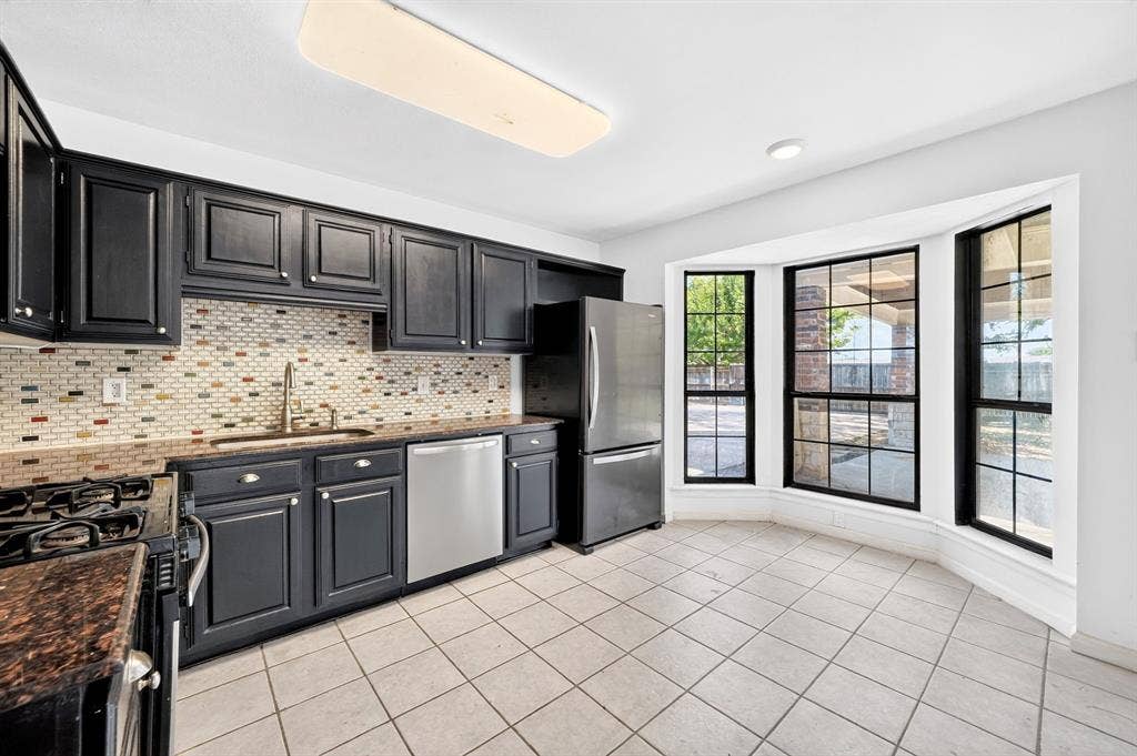 Kitchen featuring dark-finish shaker cabinetry, stone countertops, a multi-tone mosaic tile backsplash, stainless steel appliances, and a bay window