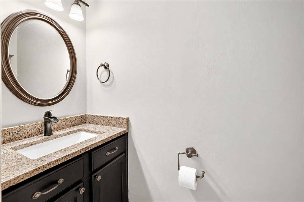 Bathroom vanity featuring dark-finished cabinetry, a stone-composite countertop, a drop-in rectangular sink, an oval wall-mounted mirror, and oil-rubbed bronze hardware