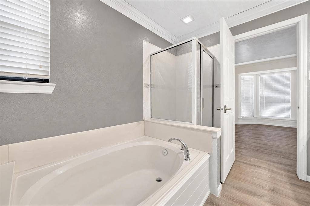 Soaking tub with chrome faucet, glass-enclosed shower, crown molding, and wood-finish flooring