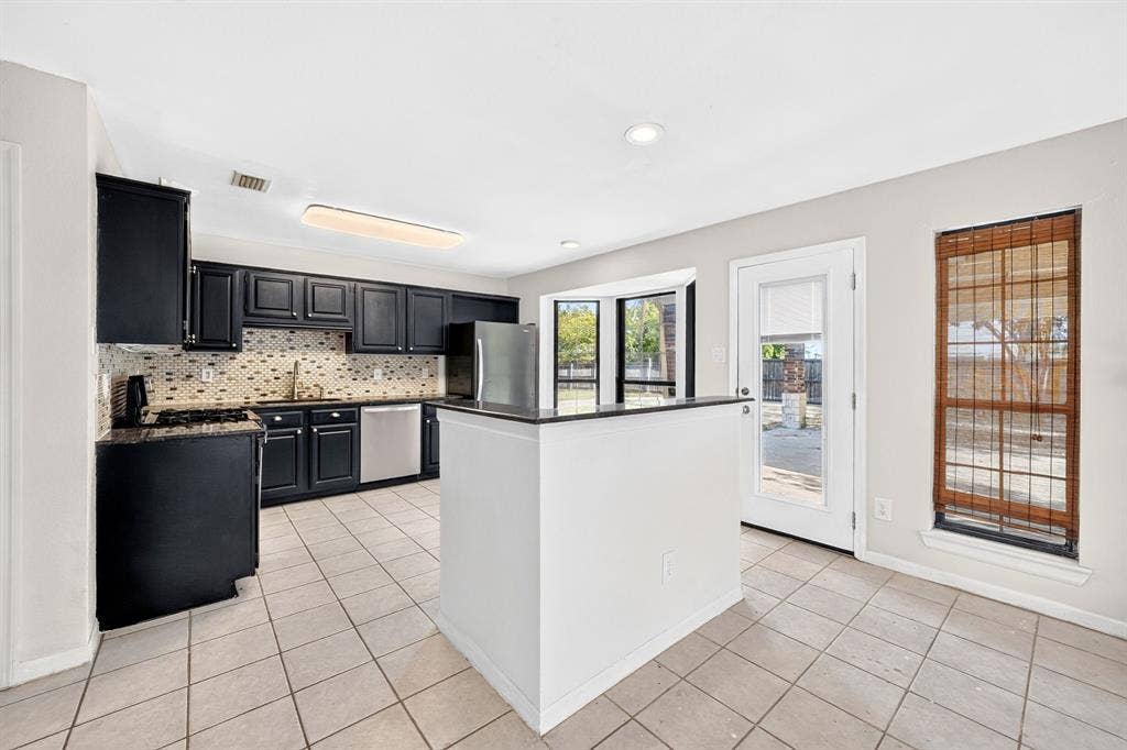 Kitchen featuring black cabinetry, a central island with a stone countertop, tile flooring, a mosaic tile backsplash, and stainless steel appliances