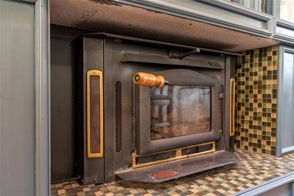 230 Bushwhacker Drive, Mabank, TX 75156 Wood-burning stove insert featuring a matte black finish, brass accents, a glass viewing panel, and a wood-toned handle, framed by mosaic tile surfacing and custom cabinetry