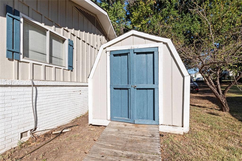 230 Bushwhacker Drive, Mabank, TX 75156 Exterior featuring a gambrel-roof storage shed with double doors, wood-sided home exterior with painted brick accents, and blue shutter detailing