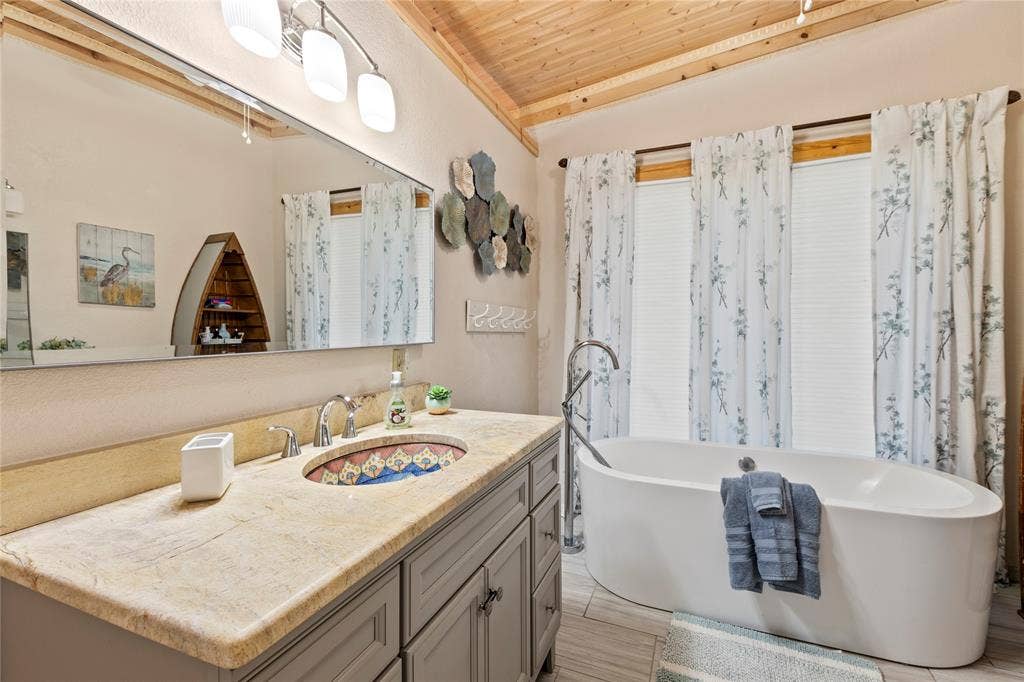 230 Bushwhacker Drive, Mabank, TX 75156 Freestanding soaking tub with a floor-mounted faucet, stone countertop with an integrated patterned basin, grey cabinetry, and wood-paneled ceiling