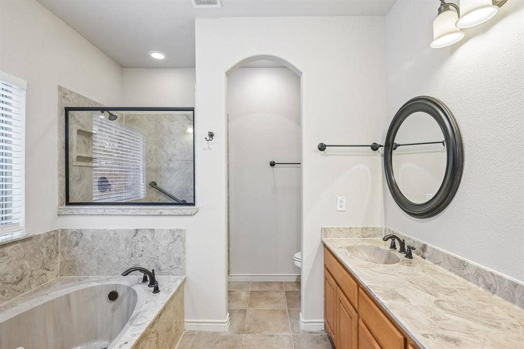 Soaking tub with stone-look surround, walk-in shower with glass enclosure, single vanity with stone-look countertop, integrated sink, and bronze-finish fixtures