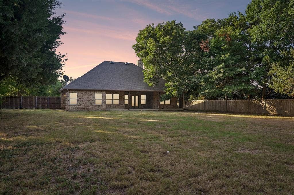 Expansive lawn with mature trees and a privacy fence, showcasing a brick exterior with multiple windows and a covered patio