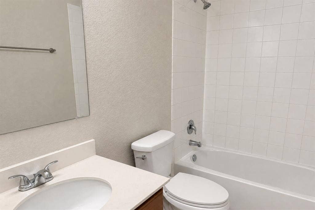 Bathroom featuring a vanity with an integrated sink and neutral countertop, wall-mounted mirror, white toilet, and a tub-shower combination with white tile surround