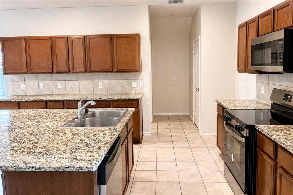 Kitchen featuring an island with a double-basin stainless steel sink, stone-finish countertops, tile backsplash, wood-finish cabinetry, and stainless steel built-in appliances
