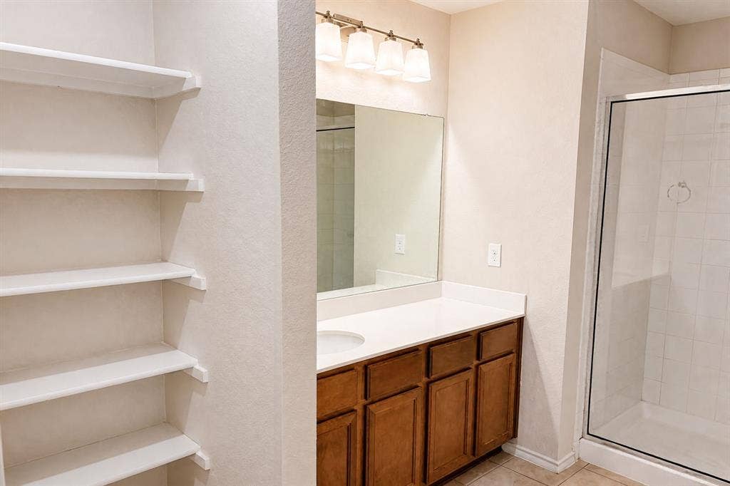 Bathroom featuring stained wood cabinetry, white countertop, integrated shelving, four-light vanity fixture, and a glass-enclosed shower with tiled surround