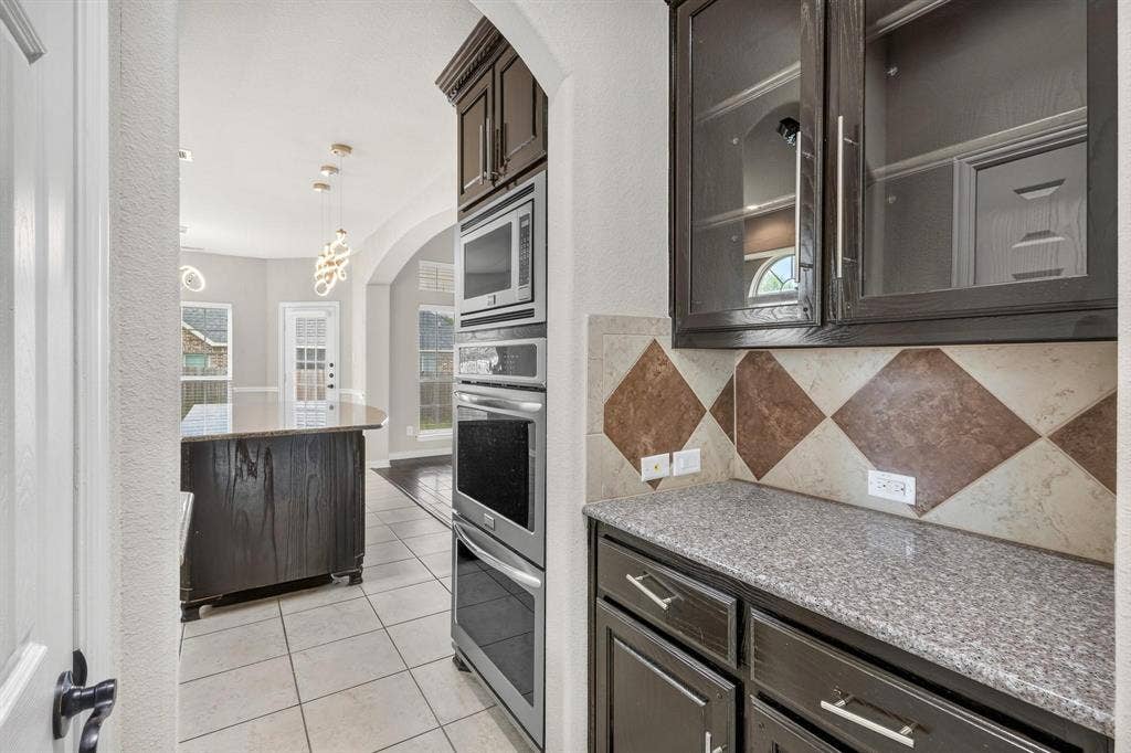 206 White Rock Court, Ovilla, TX 75154 Kitchen featuring dark wood cabinetry, built-in stainless steel double ovens and microwave, speckled stone-finish countertops, decorative tile backsplash, and ceramic tile flooring