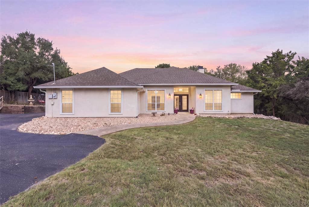 5030 Bosque Ridge Road, Crawford, TX 76638 Single-story stucco exterior featuring a multi-gabled roofline, asphalt driveway, stone landscaping, and grid-pattern windows