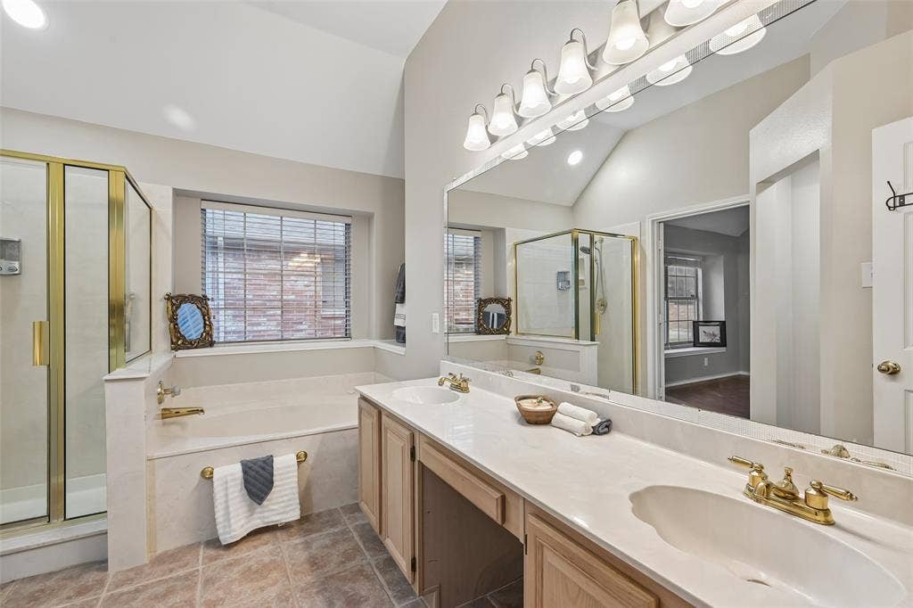 3837 Pine Valley Drive, Plano, TX 75025 Bathroom featuring dual-sink vanity with light-tone wood cabinetry, solid surface countertop, brass-finish fixtures, framed glass-enclosed shower, and built-in soaking tub