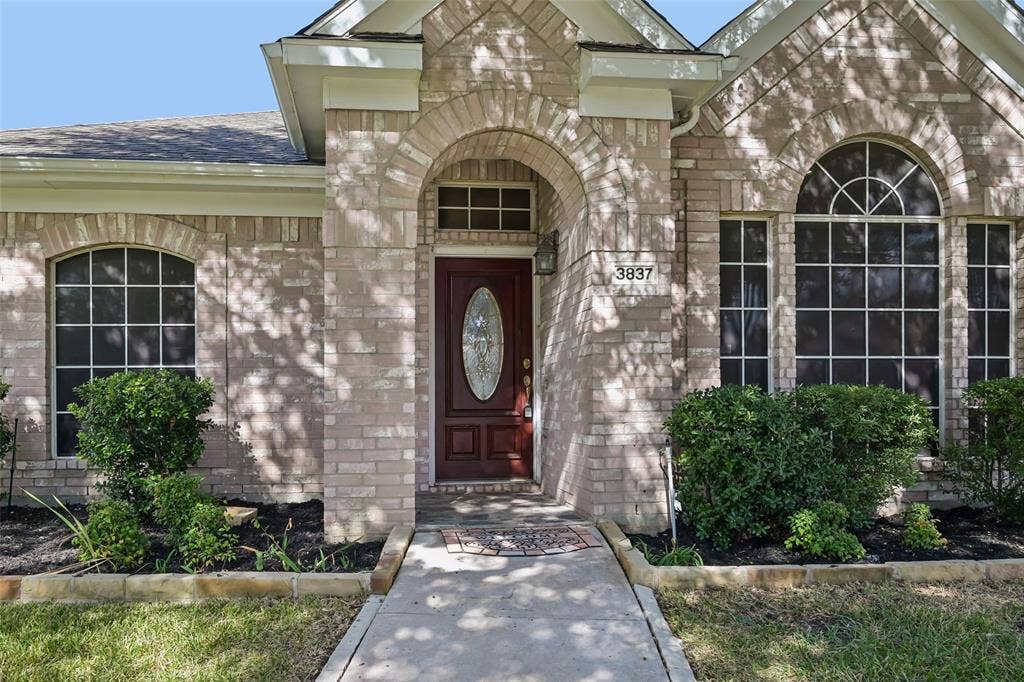 3837 Pine Valley Drive, Plano, TX 75025 Brick facade entry featuring an arched stone portico, a wood-finish front door with an oval glass insert, a transom window, and expansive grid-pane windows