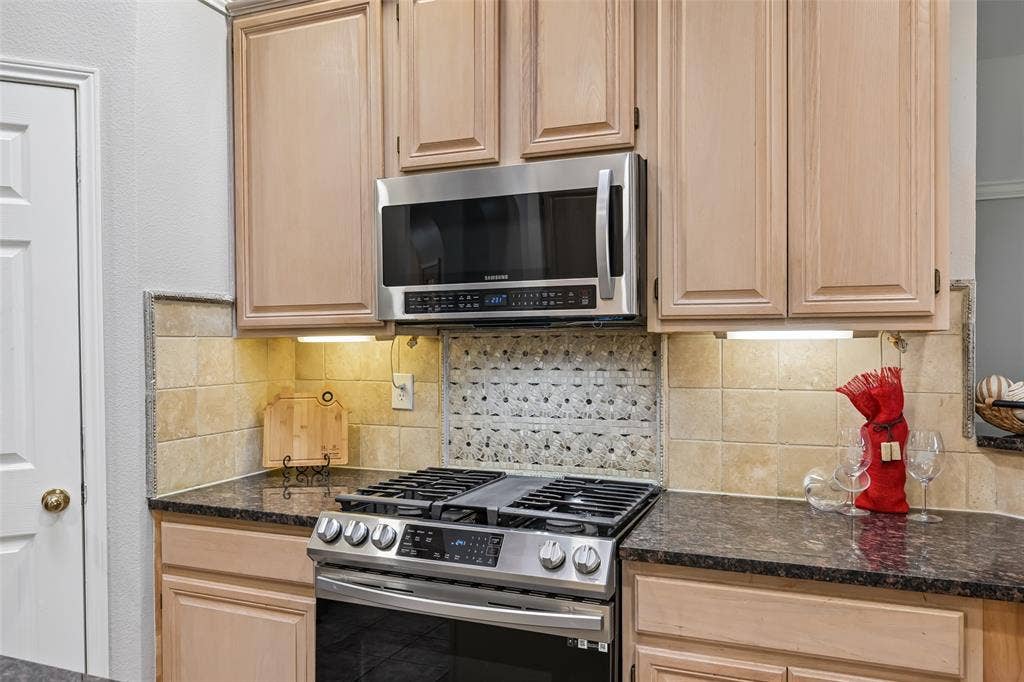 3837 Pine Valley Drive, Plano, TX 75025 Light-toned wood cabinetry, stainless steel gas range, stainless steel microwave, stone-finish countertops, and decorative tile backsplash