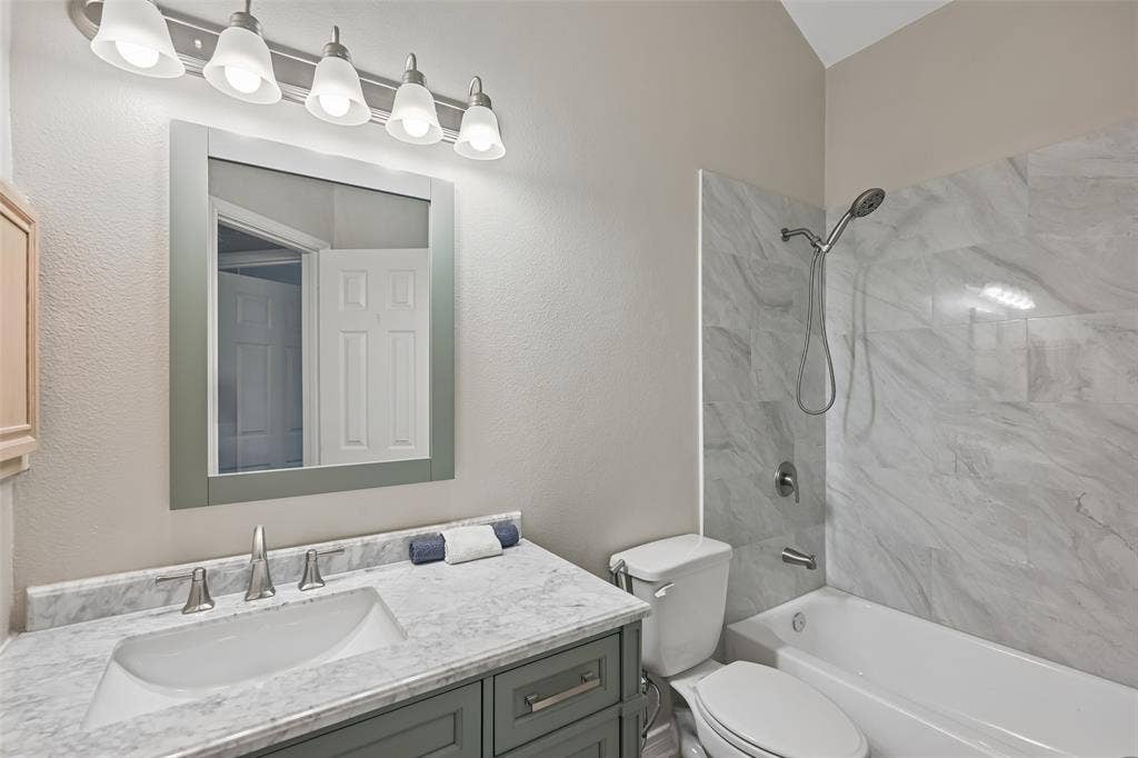 3837 Pine Valley Drive, Plano, TX 75025 Bathroom vanity featuring a stone-look countertop with an integrated rectangular sink, five-light fixture, framed mirror, and tiled shower surround