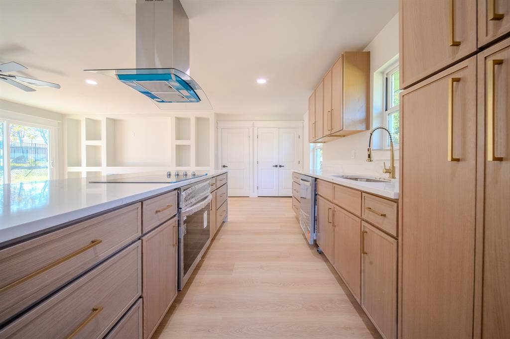 Kitchen featuring light wood-finish cabinetry, white countertops, a stainless steel range with a glass-and-steel vent hood, brass-tone hardware, and wood-finish flooring