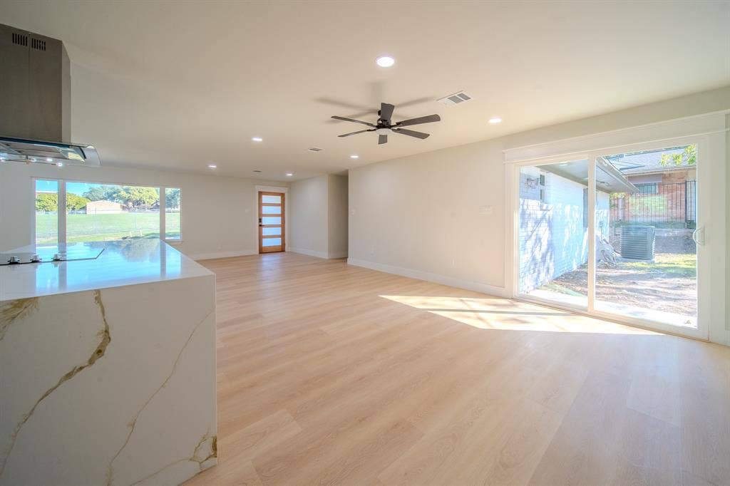 Open-concept interior featuring light wood-finish flooring, recessed lighting, a kitchen island with a stone-finish waterfall countertop, a stainless steel range hood, and sliding glass doors