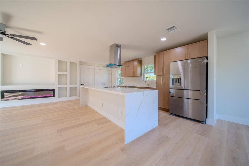 Kitchen featuring an oversized stone-finish island with waterfall edge, light-tone wood-finish cabinetry, stainless steel appliances, and a built-in electric fireplace with recessed shelving