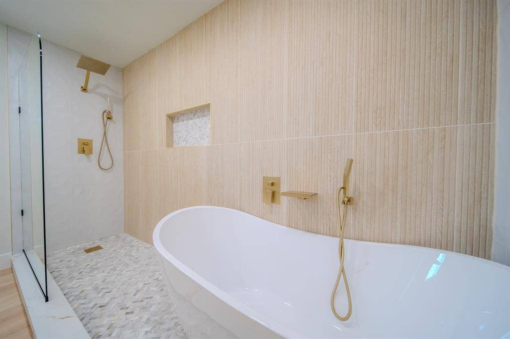 Freestanding soaking tub with gold-tone fixtures, walk-in shower with glass enclosure, vertically textured wall tiling, herringbone-patterned floor tile, and integrated wall niche