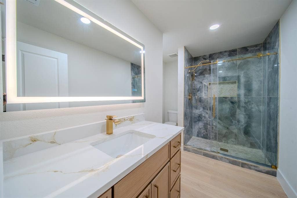 Modern vanity with gold-tone hardware and quartz countertop featuring a rectangular under-mount sink, integrated LED mirror, and walk-in shower with stone-tile surround and gold-tone fixtures