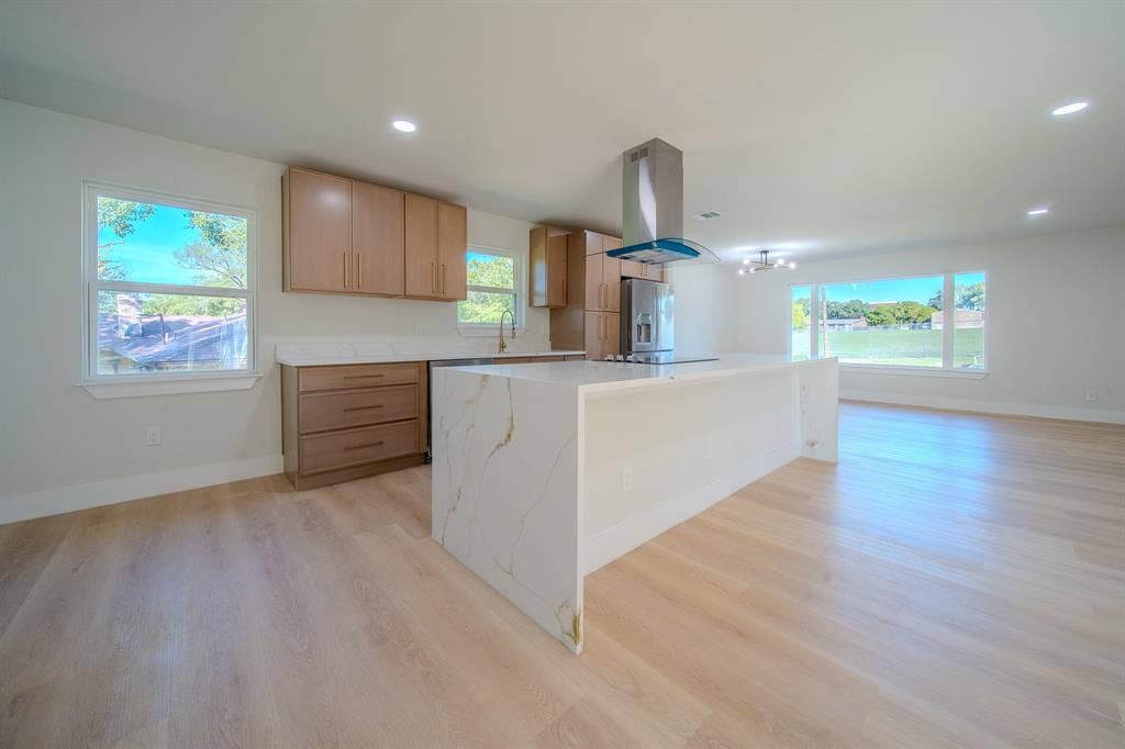 Kitchen featuring wood-finish flooring, a waterfall-edge island with stone-finish countertops, stainless steel chimney-style range hood, and flat-panel cabinetry