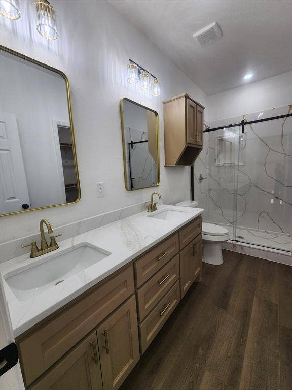 Double vanity with white stone-surface countertop, integrated rectangular sinks, brushed gold-tone faucets, wood-finish cabinetry, and glass-enclosed shower with wall-to-wall large-format tile surround
