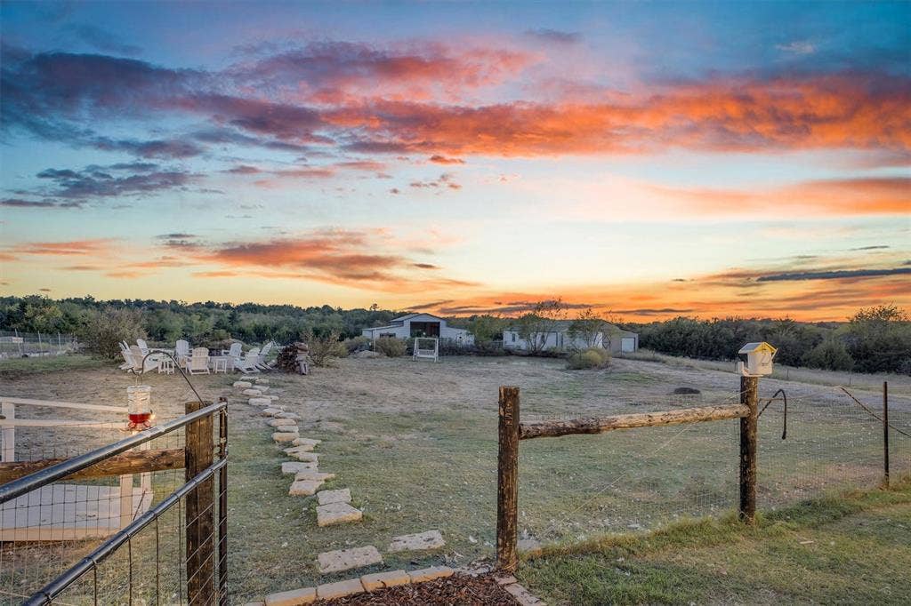 9880 County Road 466, Princeton, TX 75407 Expansive grounds featuring a stone paver path, wooden post-and-rail fencing, and outbuildings against a prominent sunset horizon