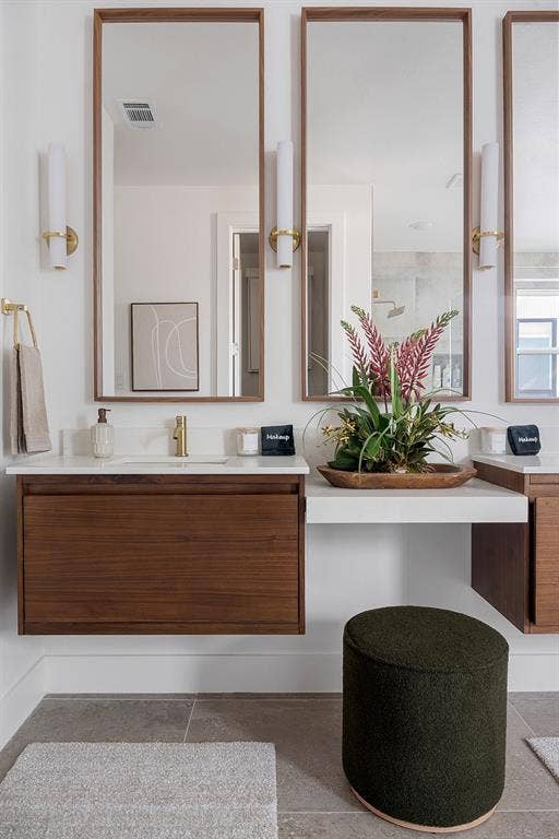Floating wood-finish vanity with a stone countertop, rectangular undermount sink, brushed gold faucet, wall-mounted cylinder sconce lighting, and minimalist wood-framed mirrors