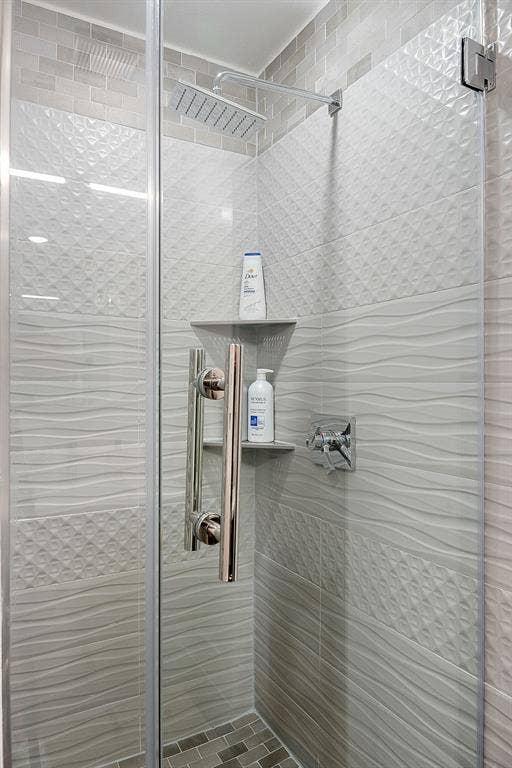 Walk-in shower featuring a frameless glass door, rain-style showerhead, chrome fixture hardware, tiered corner shelving, and multi-textured tile wall detailing