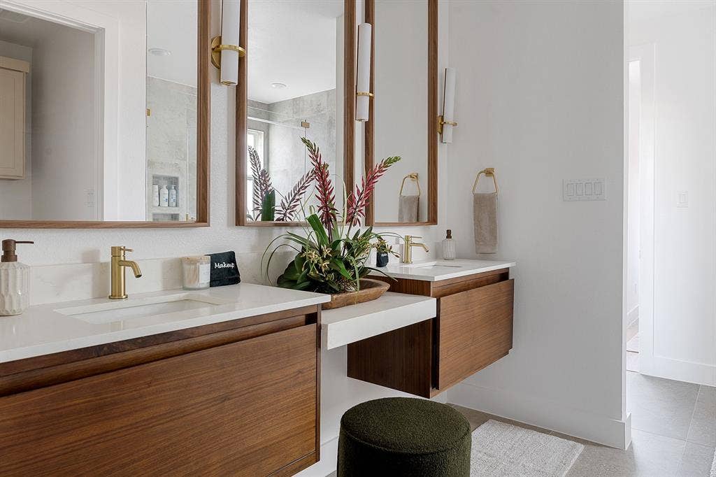 Floating dual-vanity with wood cabinetry, white stone countertops, integrated sinks, brass-toned fixtures, and vertical-frame mirrors with matching sconces