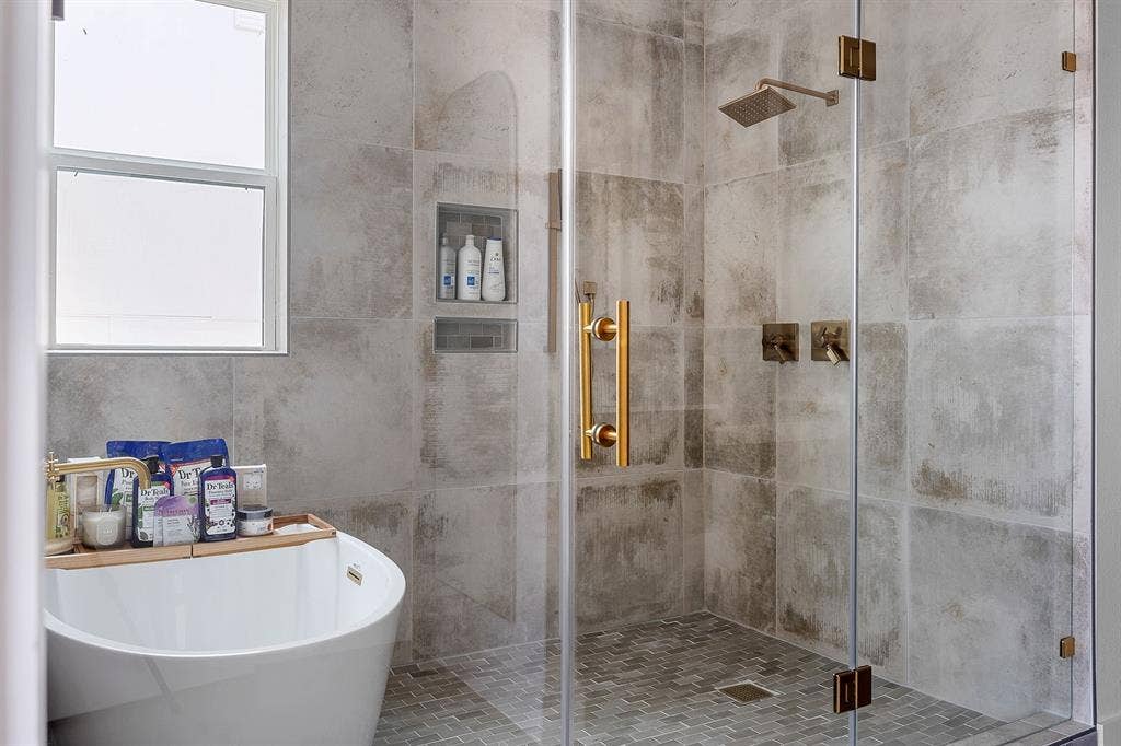 Walk-in shower featuring large-format neutral wall tile, mosaic tile flooring, glass enclosure, brass-finish fixtures, and integrated wall niches