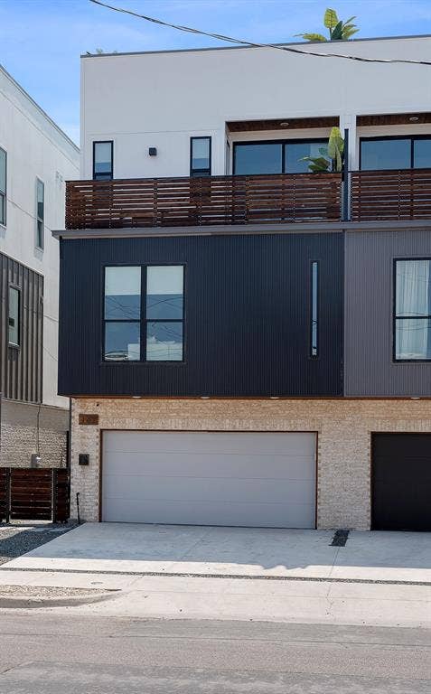 Modern multi-level exterior featuring white stucco, black vertical siding, light-toned stone masonry, horizontal wood-slat balcony railings, and a concrete driveway