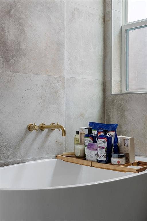 Freestanding soaking tub featuring large-format stone-finish wall tile, brass wall-mounted faucet hardware, and a window
