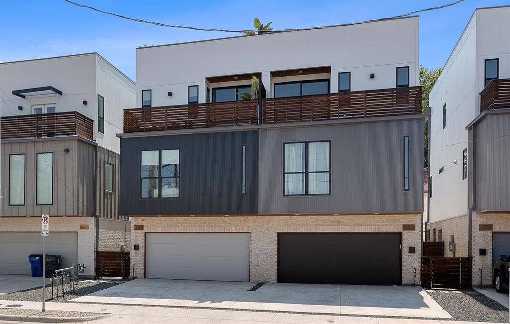 Modern multi-story exterior featuring a stone veneer base, mixed-material metal siding, wood-slat balcony railings, and dual garage access
