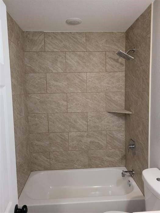 Bathtub and shower combination featuring large-format neutral-toned tile surround, built-in corner shelving, chrome rainfall showerhead, and matching chrome faucet fixture