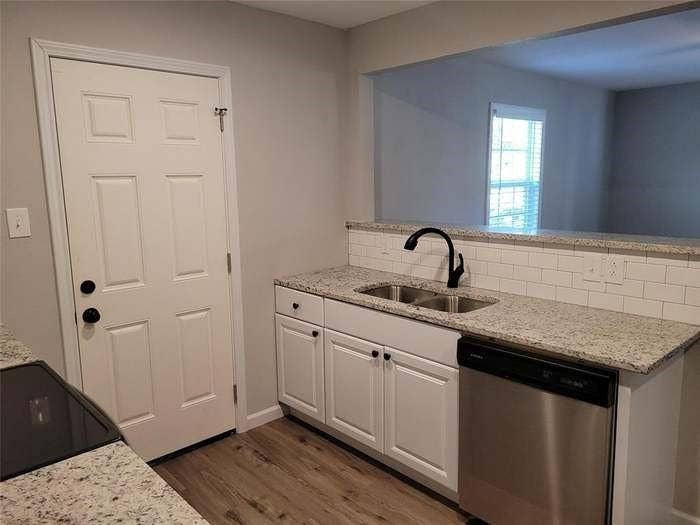 Kitchen area featuring white shaker-style cabinetry, stone-surface countertops, subway tile backsplash, stainless steel dishwasher, and wood-finish flooring