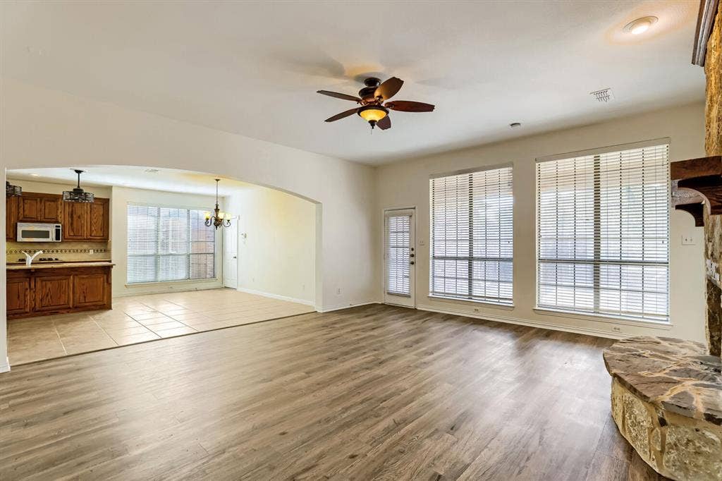 7013 Salem Court, Rowlett, TX 75089 Open-concept interior featuring wood-finish flooring, a stone-accented fireplace hearth, large windows with horizontal blinds, a vaulted ceiling with a central fan, and ceramic tile flooring in the kitchen area