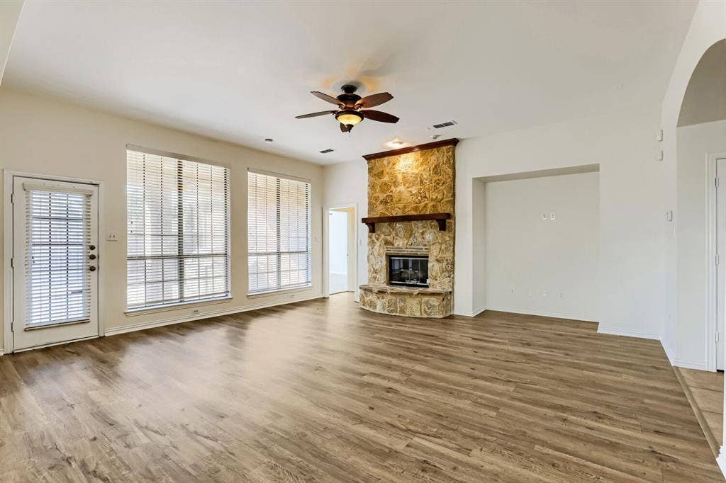 7013 Salem Court, Rowlett, TX 75089 Living area featuring wood-finish flooring, floor-to-ceiling stone fireplace with a timber mantel, high ceilings, ceiling fan, and large windows with integrated blinds