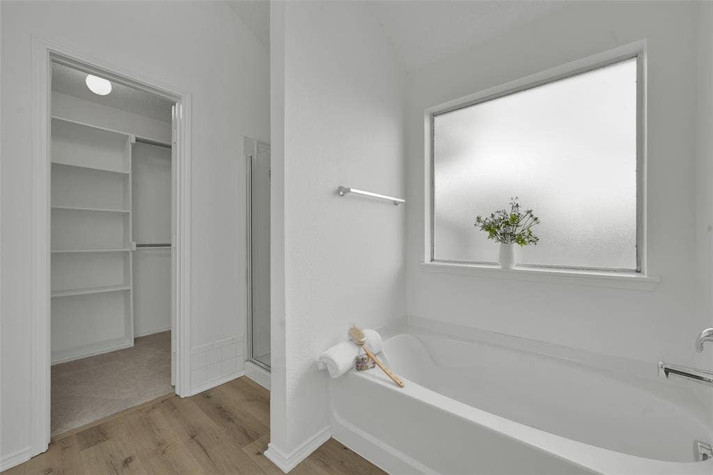 Deep soaking tub with chrome fixtures, frosted privacy window, walk-in closet with built-in shelving, and wood-finish flooring