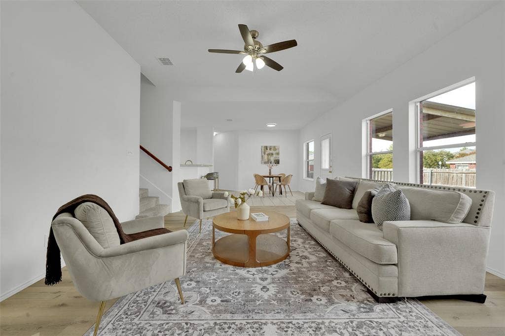 Open-concept interior featuring wood-finish flooring, neutral walls, multiple windows providing natural light, and a ceiling fan with integrated lighting