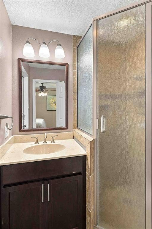 18240 Midway Road, #1706, Dallas, TX 75287 Bathroom featuring a dark wood-finish vanity, light-toned countertop with integrated oval basin, three-light wall fixture, framed mirror, and walk-in shower with textured glass enclosure