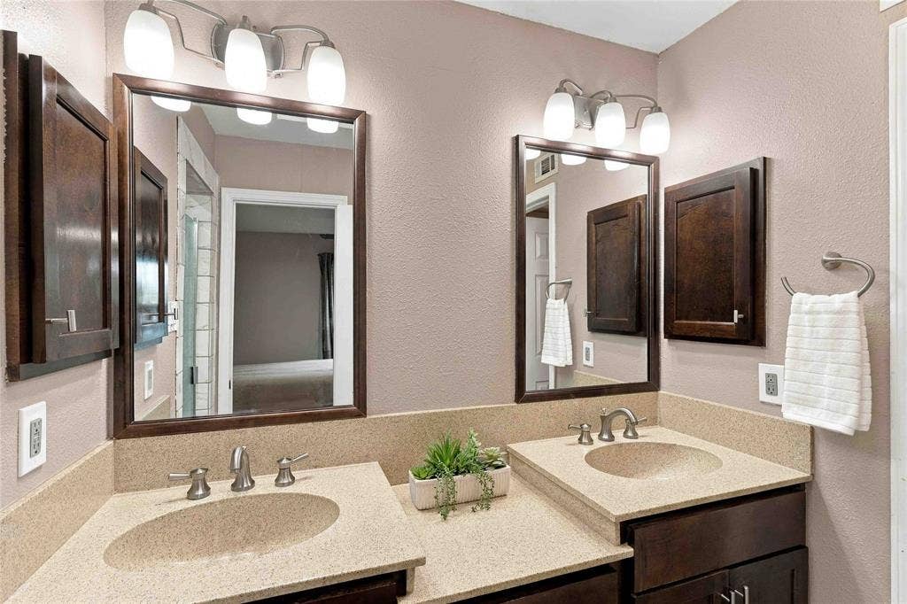 18240 Midway Road, #1706, Dallas, TX 75287 Dual-sink vanity featuring stone-composite countertops, dark wood-finish cabinetry, matching framed mirrors, and wall-mounted tri-light fixtures