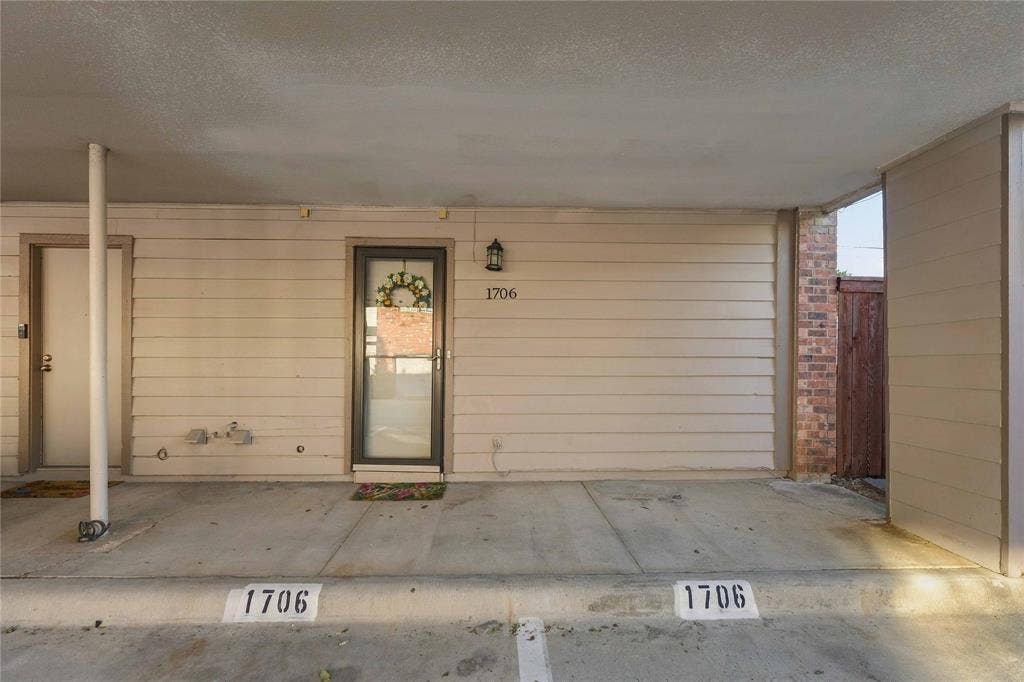 18240 Midway Road, #1706, Dallas, TX 75287 Covered carport entrance featuring horizontal siding, concrete flooring, exterior wall-mounted lighting, and a glass-paneled entry door