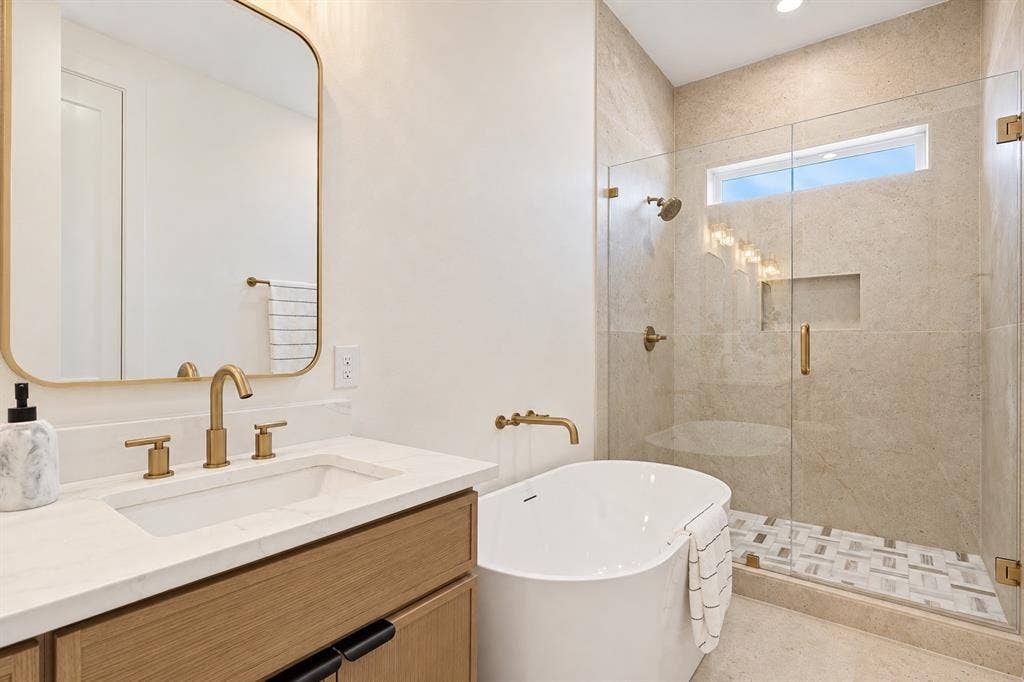 Freestanding tub with wall-mounted faucet, glass-enclosed shower with contrasting tile floor, single vanity with wood-finish cabinetry, and gold-tone fixtures throughout