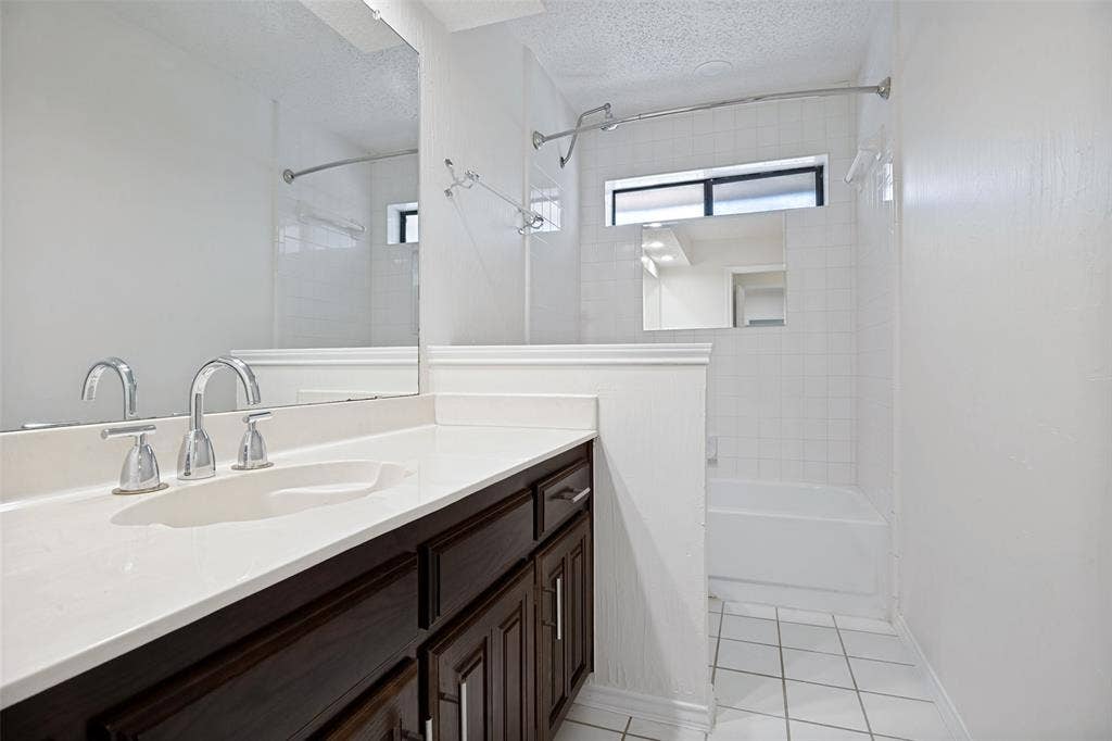 6627 Sawmill Road, Dallas, TX 75252 Bathroom featuring dark-stained cabinetry, a white countertop with an integrated basin, a polished chrome dual-handle faucet, a full-width mirror, and a tile-surround bathtub