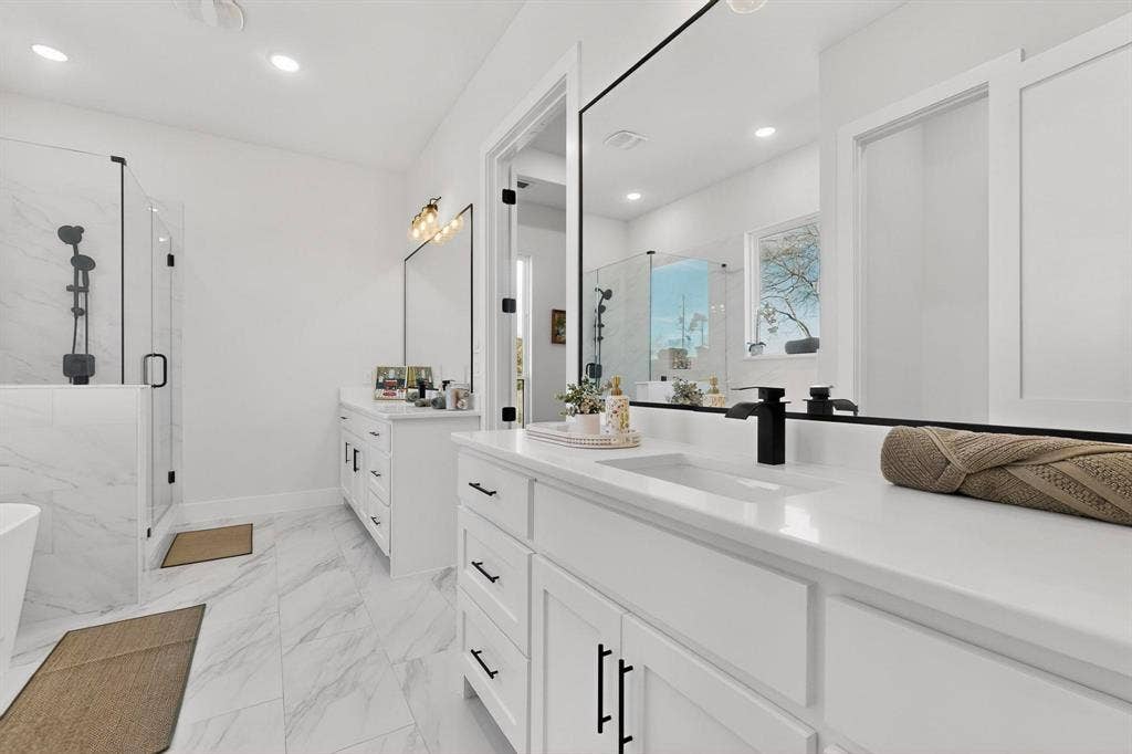 Bathroom featuring dual white cabinetry with matte black hardware, stone-finish countertops, expansive wall-to-wall mirrors, and a glass-enclosed shower with large-format tile surrounds