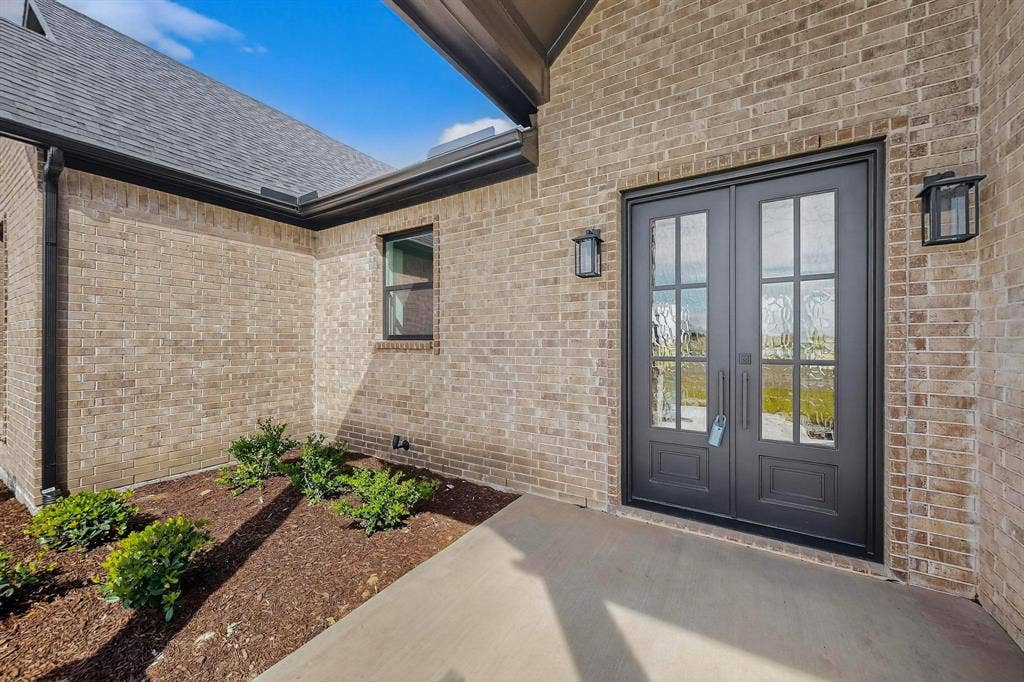 Covered entryway featuring light-toned brick facade, double front doors with glass panels, dark metal framing, and wall-mounted exterior lanterns