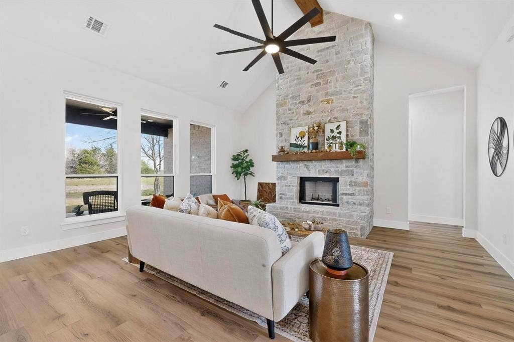 Vaulted ceiling with an exposed wood beam, floor-to-ceiling stone fireplace, prominent wood-finish flooring, multi-pane floor-to-ceiling windows, and a large-format multi-blade ceiling fan
