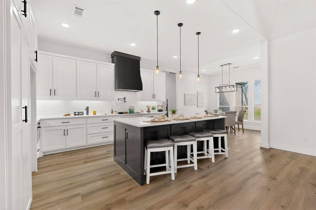 Kitchen featuring wood-finish flooring, white cabinetry, a center island with a contrasting dark base, a built-in range hood, and three pendant lights