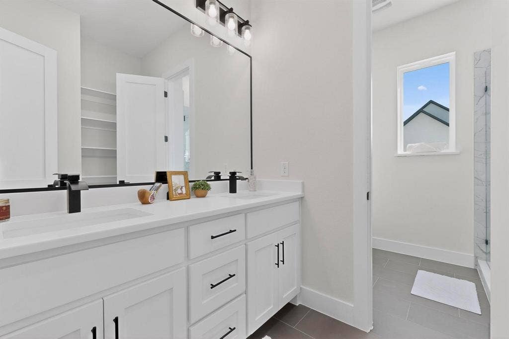 Dual-sink vanity with white cabinetry, solid surface countertop, and matte black hardware, accompanied by a large framed mirror, vanity lighting, and a glass-enclosed shower with tile flooring